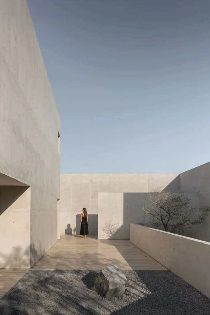 Open-air courtyard with exposed concrete volumes, gravel garden, natural boulder, long shadows, and a solitary figure in a quiet contemplative set