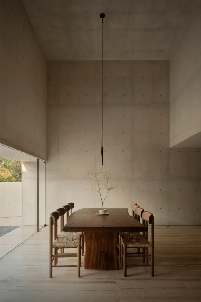 Spacious concrete living room with sculpture wooden seat, minimal furnishing, high ceilings, and a blurr figure walk through the soft lit interior