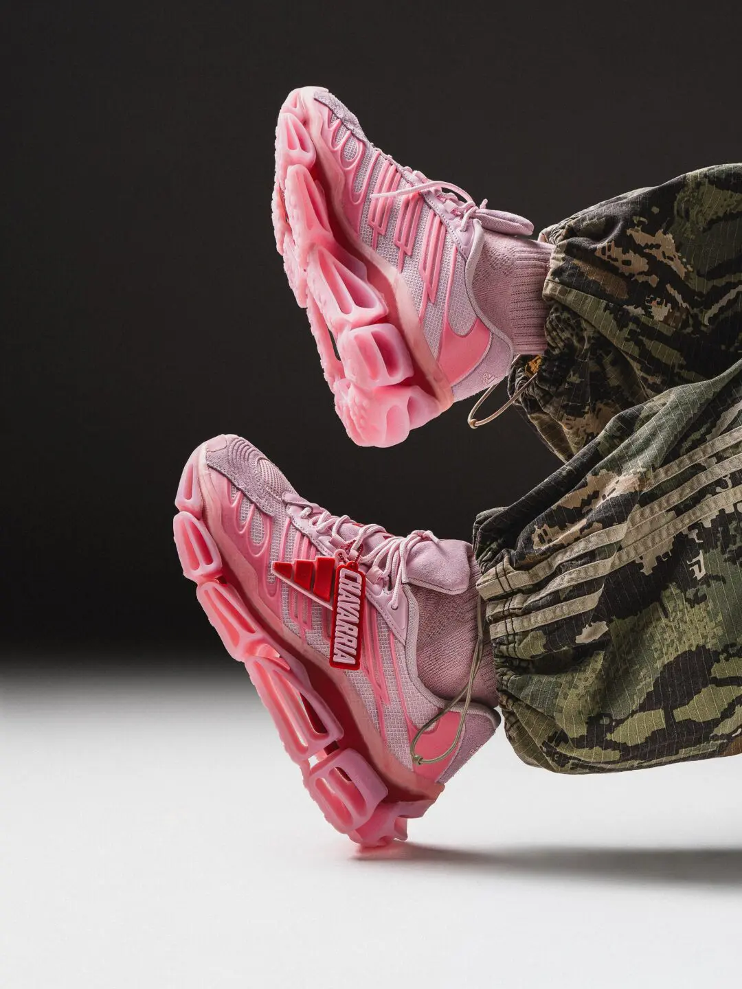 On-foot view of the adidas x Willy Chavarria pink sneaker featuring layered mesh and suede panels, oversized hollow midsole cushioning, a removable co-branded Chavarria hangtag, and camouflage track pants against a dark studio backdrop.