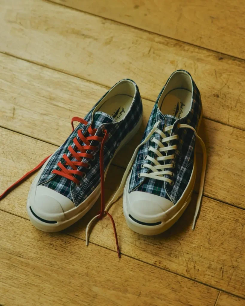Pair of Noah x Jack Purcell tartan low-top shoe on a wooden floor, style with mismatch red and white laces and off-white rubber toe cap