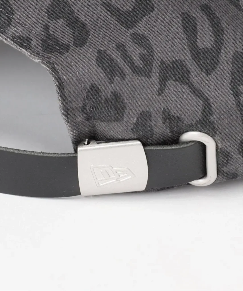 Close-up of a gray textured cap with a black strap, silver buckle bearing an embossed logo, and white loop on a light