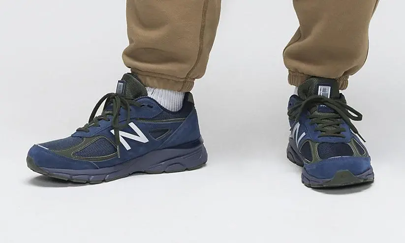 Blue And Olive New Balance Running Shoes Styled With Tan Jogger Pants Against A White Studio Background