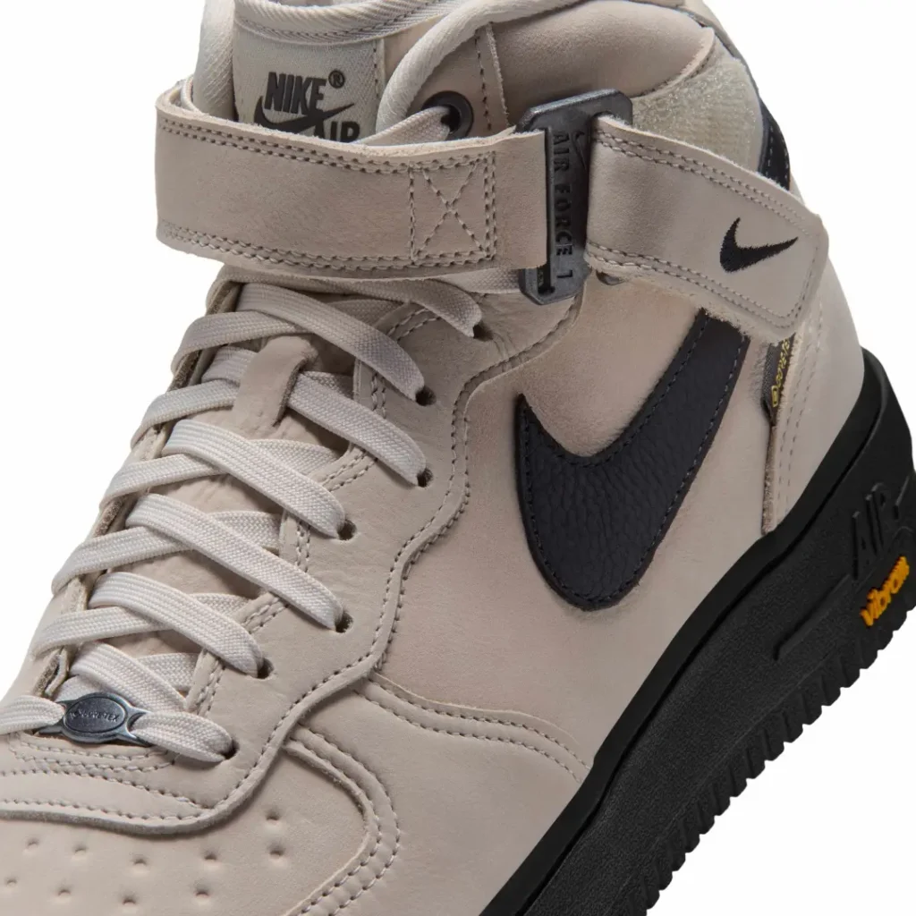 Close-up of the beige Nike Air Force 1 Mid show tonal lace, ankle strap hardware, black Swoosh and Vibram brand