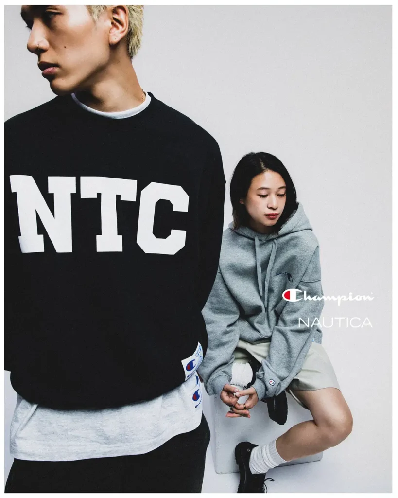 Two model wear Champion x Nautica apparel, include a black oversized NTC sweatshirt and gray Nautica hoodie, against a min white studio backdrop