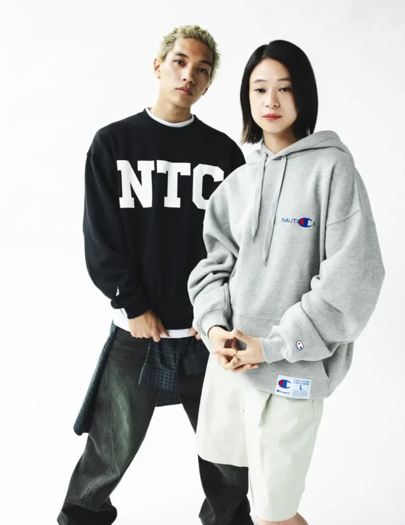 Two model pose in Champion x Nautica apparel, feature a black NTC sweatshirt and oversize gray Nautica hoodie against a bright white background