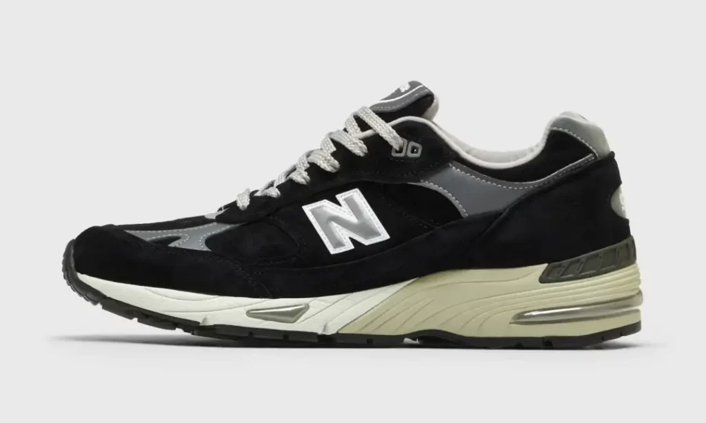Black and grey New Balance 991 shoe shown in lateral profile, featuring premium suede and mesh panels, reflective silver accents, grey laces, and a cream ABZORB midsole against a light grey background