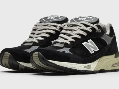 Pair of black and grey New Balance 991 shoe viewed from the front three-quarter angle, show laye suede construction, reflective details, chunky cream midsoles, and texture rubber outsole
