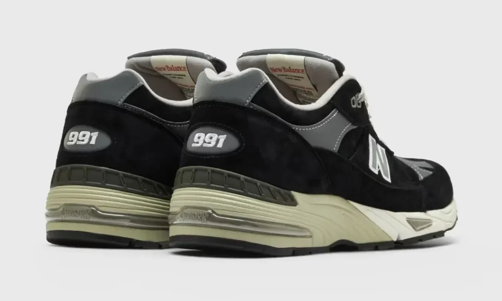 Rear three-quarter view of a pair of New Balance 991 show, featuring embroidered "991" heel branding, black suede uppers, grey leather collars, cream midsoles with visible ABZORB cushion, and durable black outsole