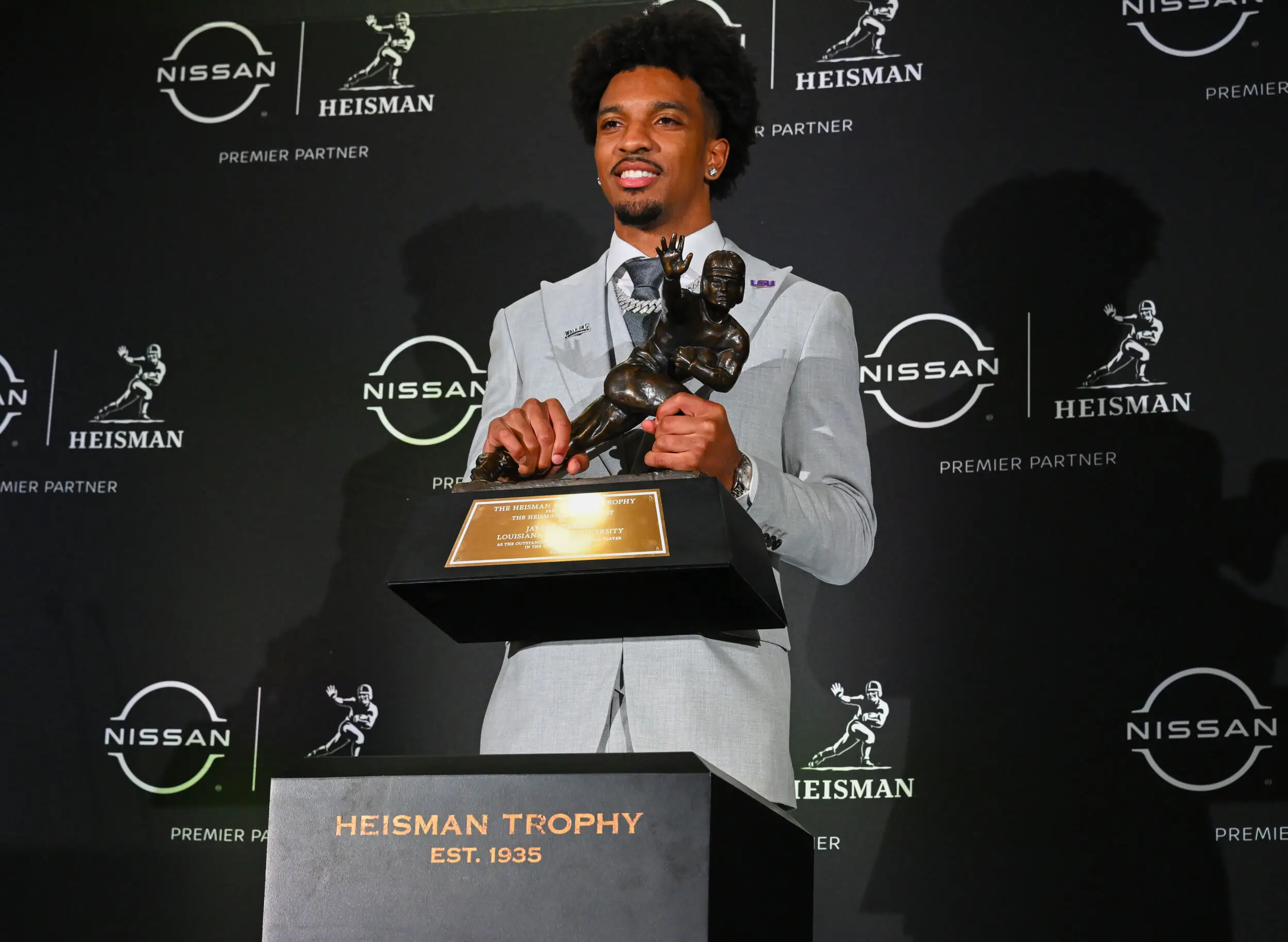 College Football Player Poses With The Heisman Trophy While Wearing A Light Gray Suit At The Heisman Trophy Ceremony, With Nissan And Heisman Branding Across The Black Backdrop.