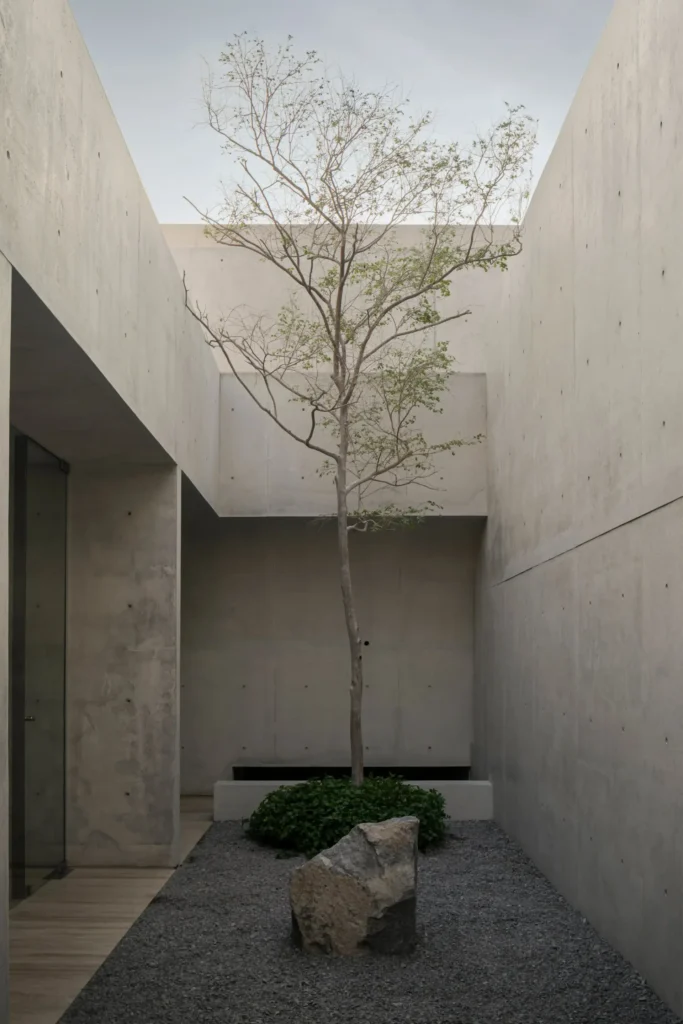 Min bedroom opening onto a private gravel courtyard through floor-to-ceil glazing, with expose concrete walls and a low platform bed