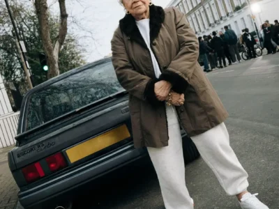 An older woman in white Reebok shoe stands beside a black Ford Escort RS Turbo on a London street