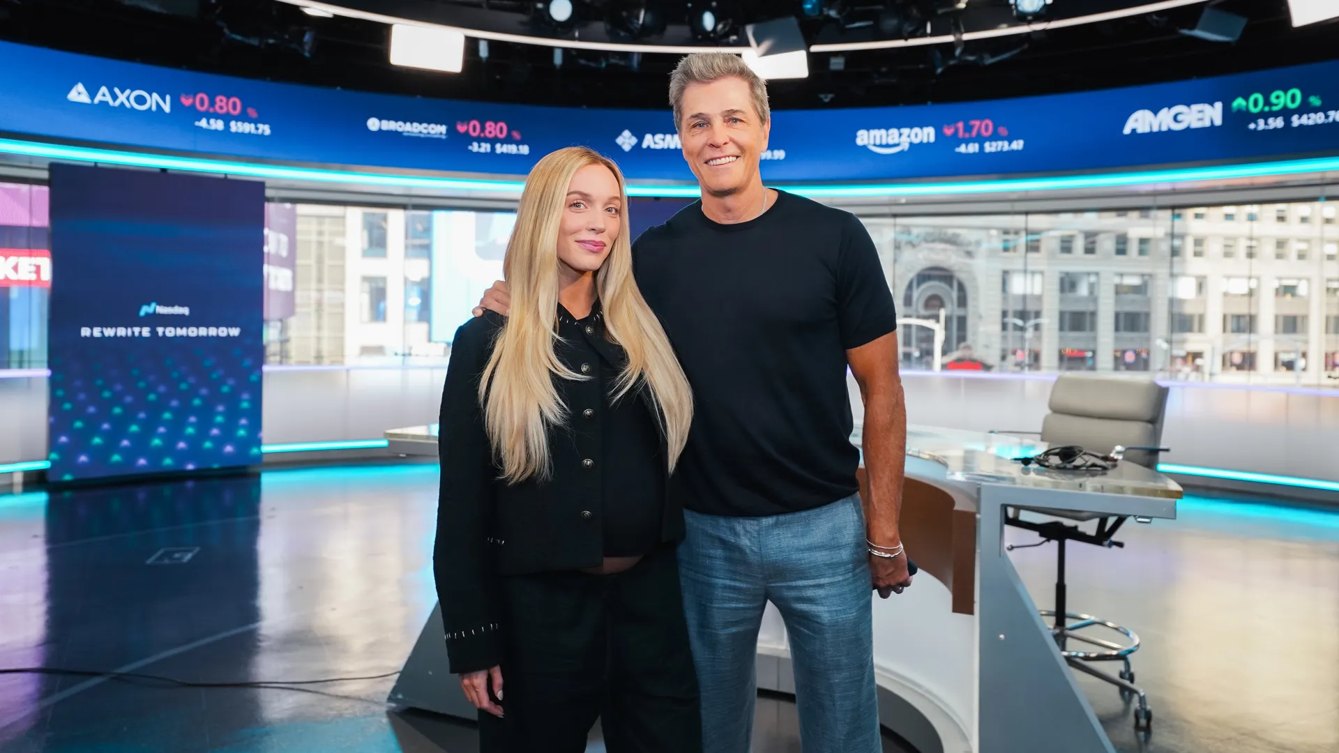 Blonde Woman And Male Television Host Posing Together Inside A Modern Financial News Studio With Market Displays In The Background