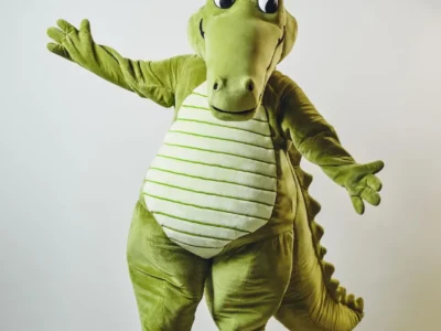 A plush lime Crocodile mascot poses with arms wide open. He wears oversize white clogs decorate with jibbitz while standing inside a min studio space