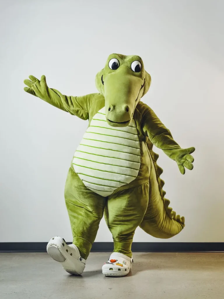 A plush lime Crocodile mascot poses with arms wide open. He wears oversize white clogs decorate with jibbitz while standing inside a min studio space