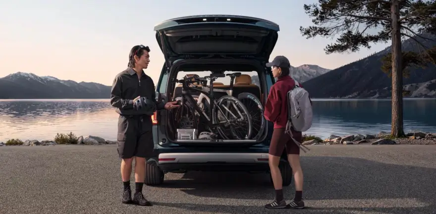Two Cyclists Standing Beside An Open SUV Loaded With Bicycles And Outdoor Gear Along A Mountain Lake
