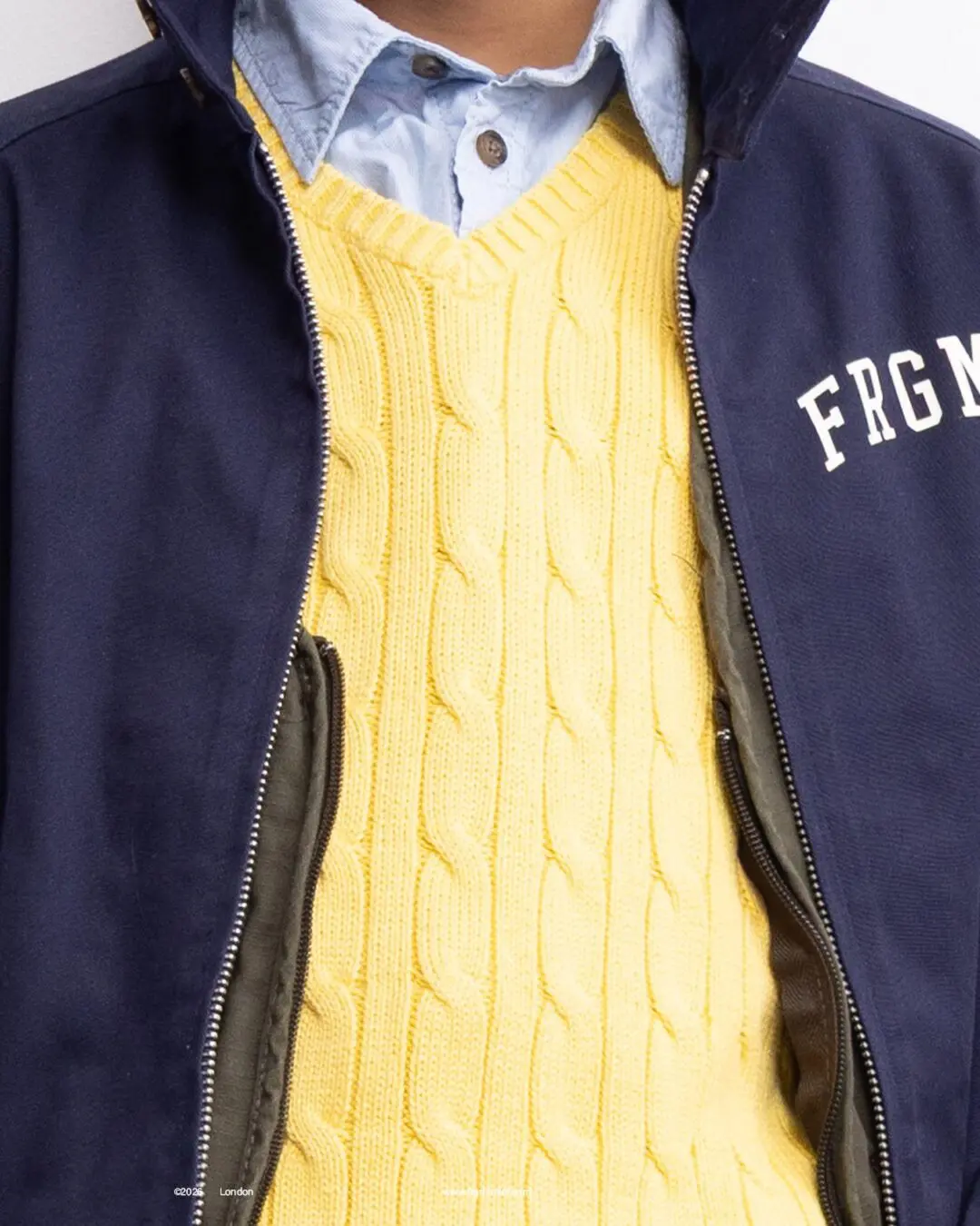 Close-up of a yellow cable-knit sweater layered under a blue collared shirt and navy jacket with white lettering