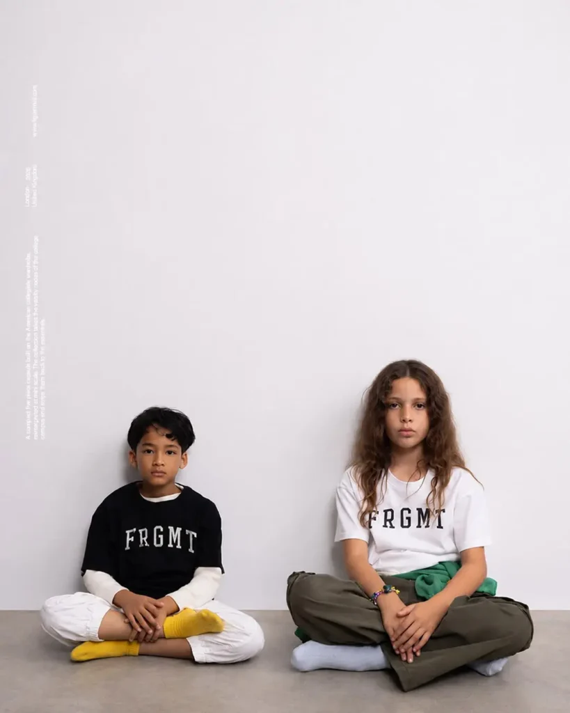Two children sitting on concrete: boy in black FRGMT tee, girl in white FRGMT tee with green pant