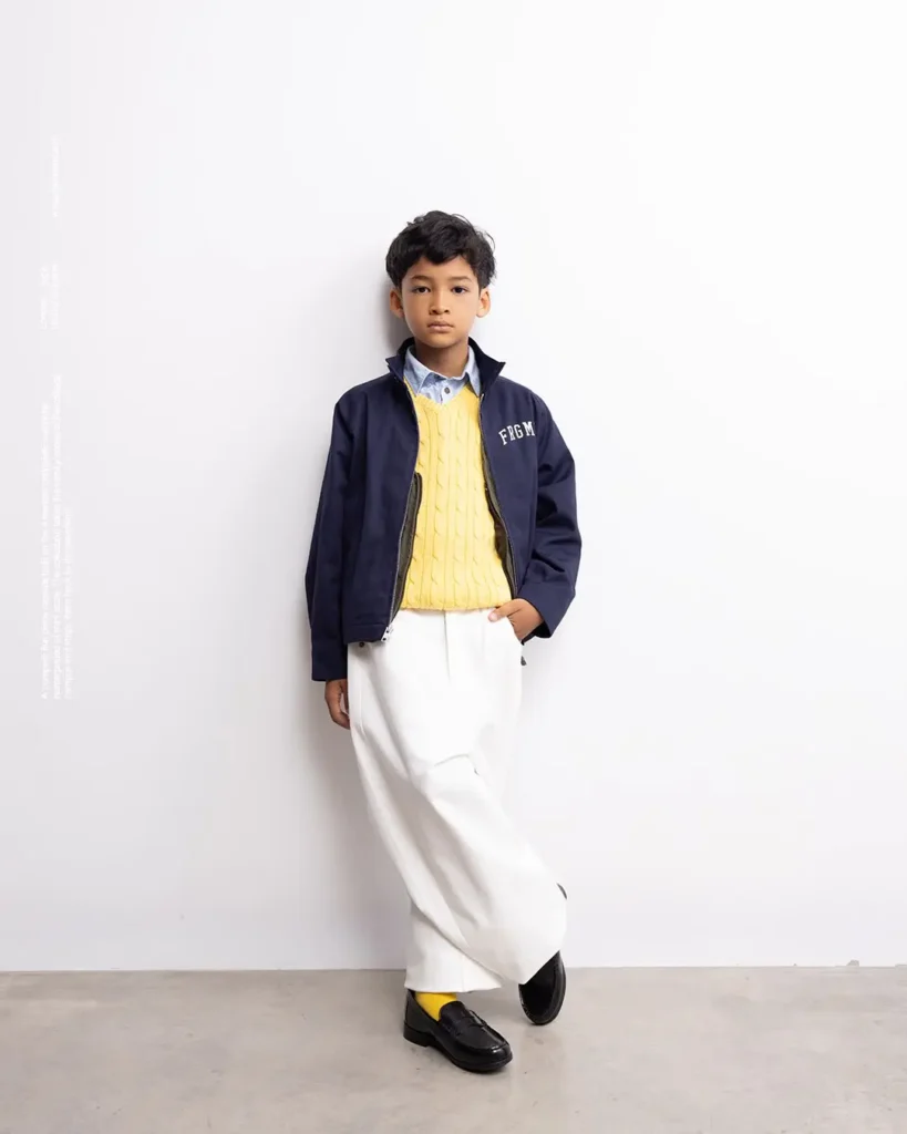 Boy in navy FRGM jacket, yellow cable-knit sweater, white trouser, and black loafer by a white wall