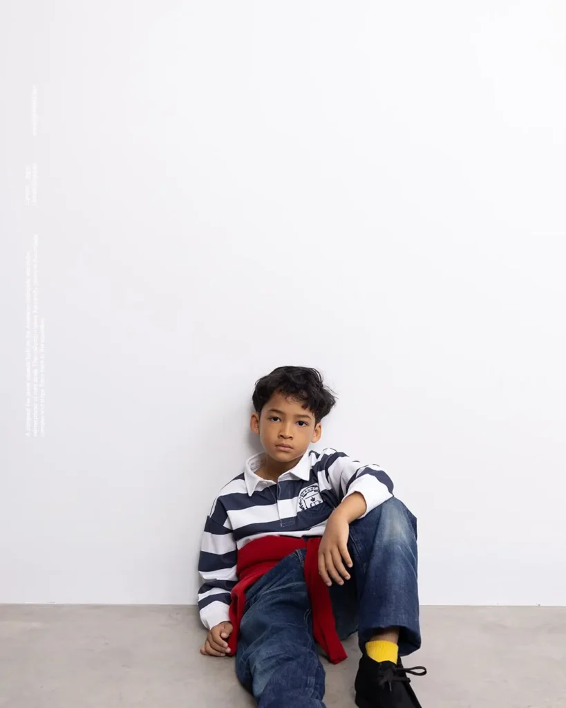 Boy sitting against a white wall in navy-white striped rugby shirt, red sweater at waist, and jean