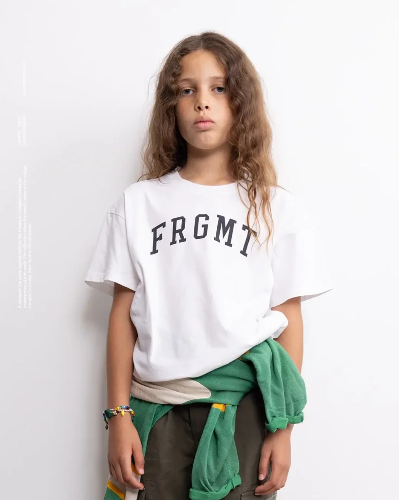 Girl in oversize white FRGMT t-shirt with green sweater tied at waist and olive pant, stood