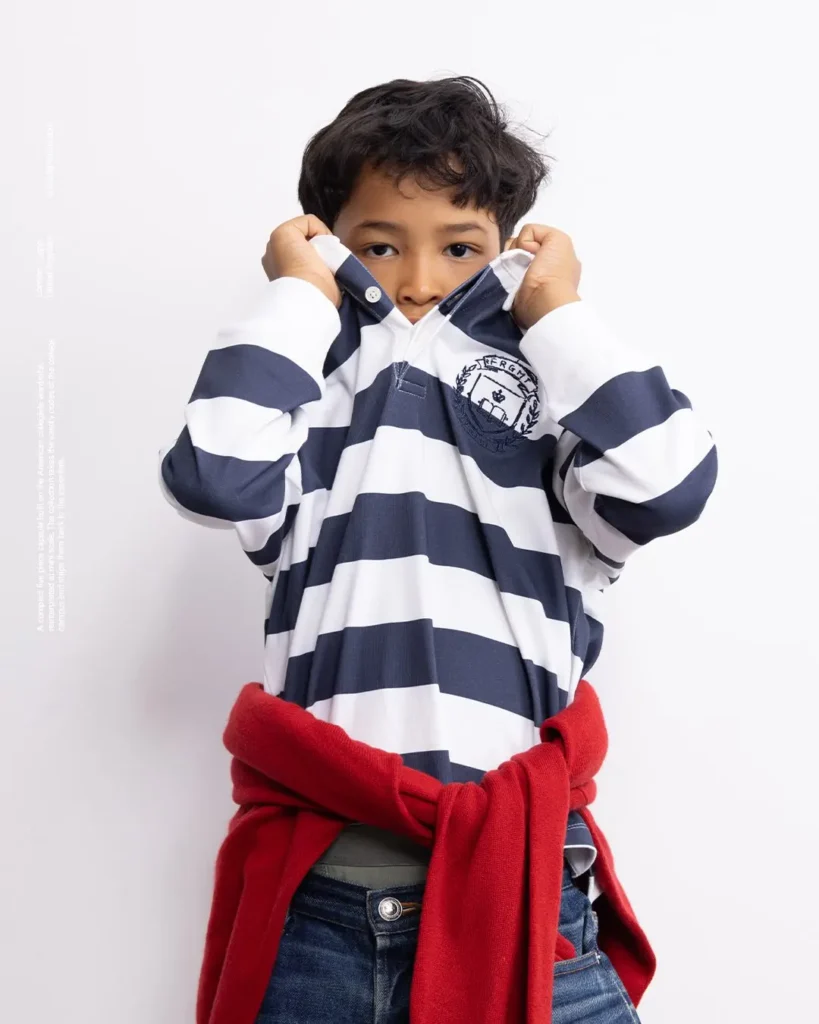 Boy pull up the collar of a navy-white striped FRGMT rugby shirt with red sweater around his waist