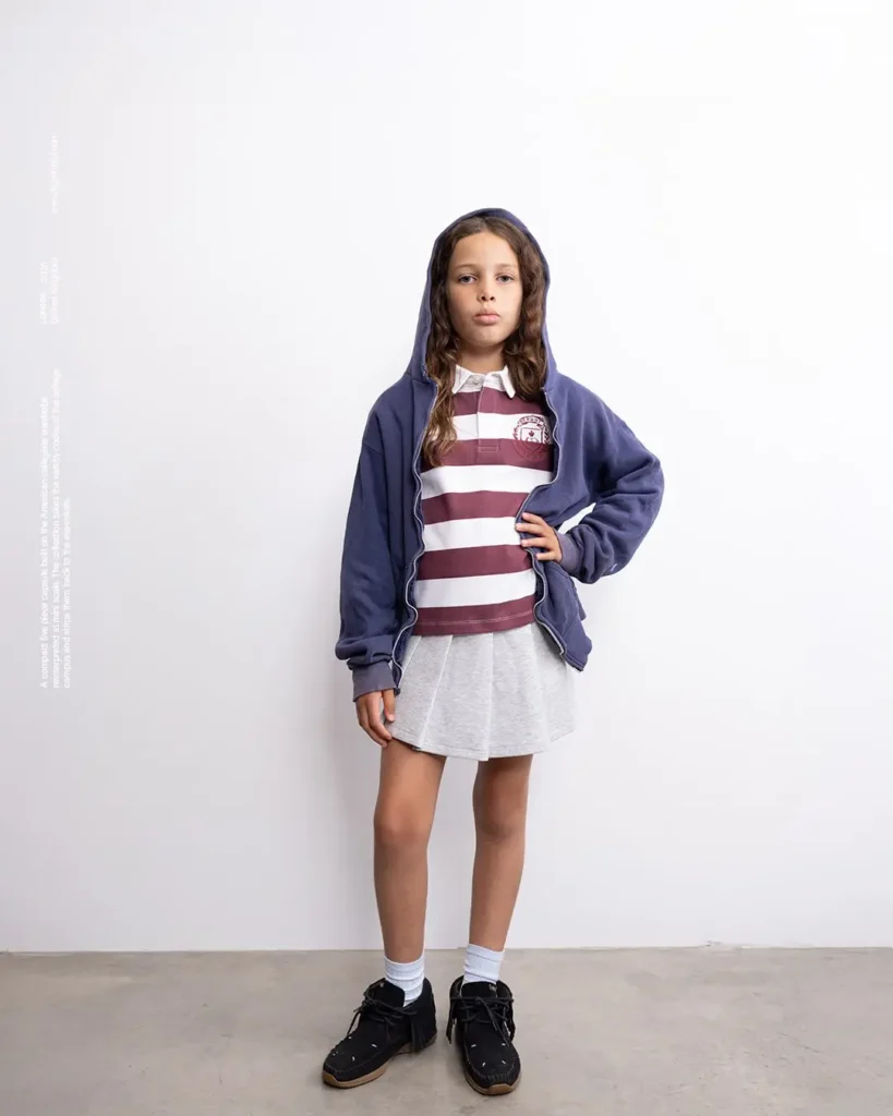 Girl in blue zip hoodie over maroon-white stripe rugby, grey skirt, and black shoe stood by a wall