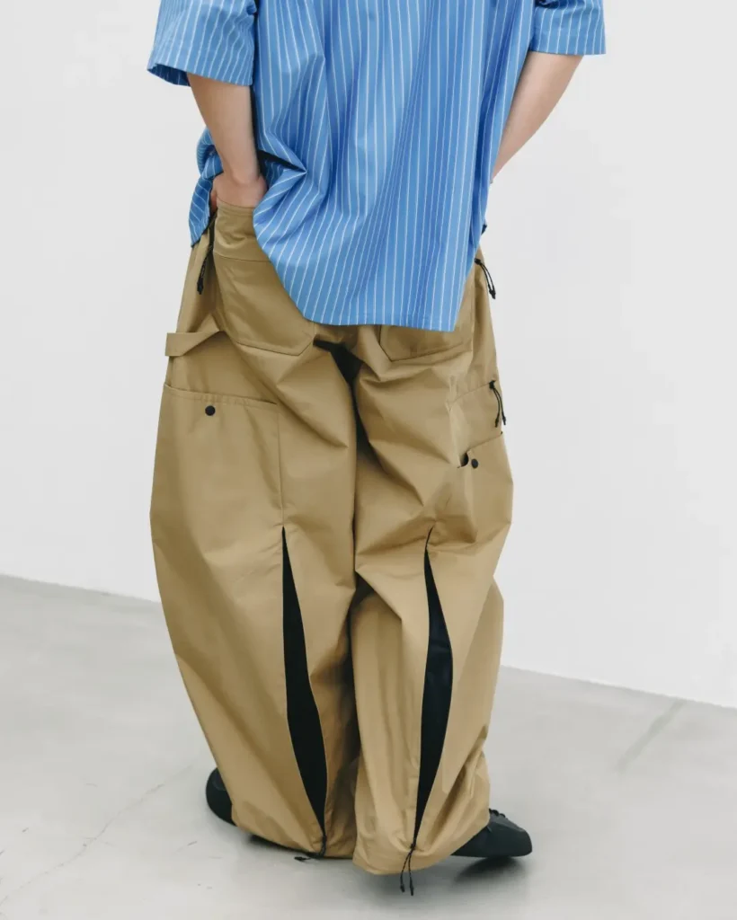 Rear view of a model wearing tan/khaki balloon-fit utility pants with hand in back pocket, showing dual leg-seam vents lined in black, paired with a blue pinstripe shir