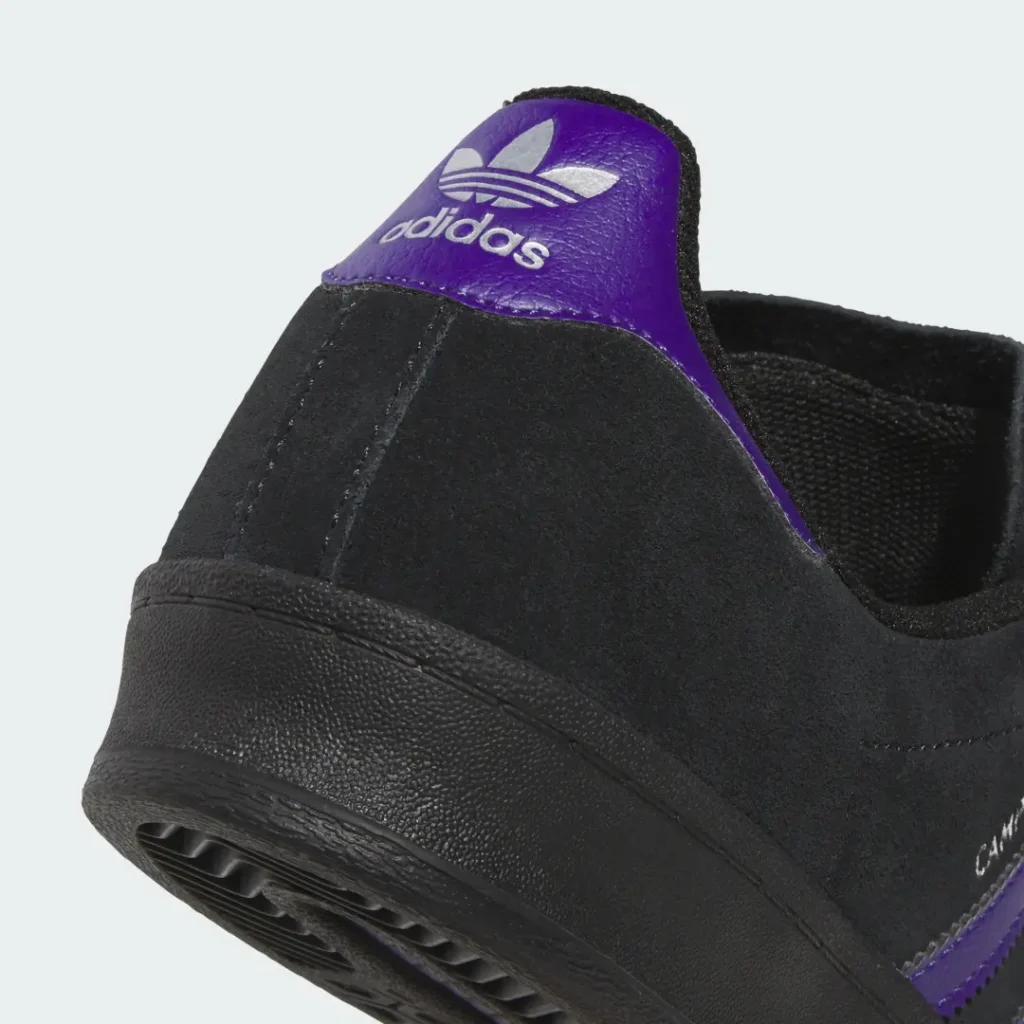 Close-up of a black sneaker heel with a purple leather collar, silver Adidas trefoil, and textured black sole on a pale