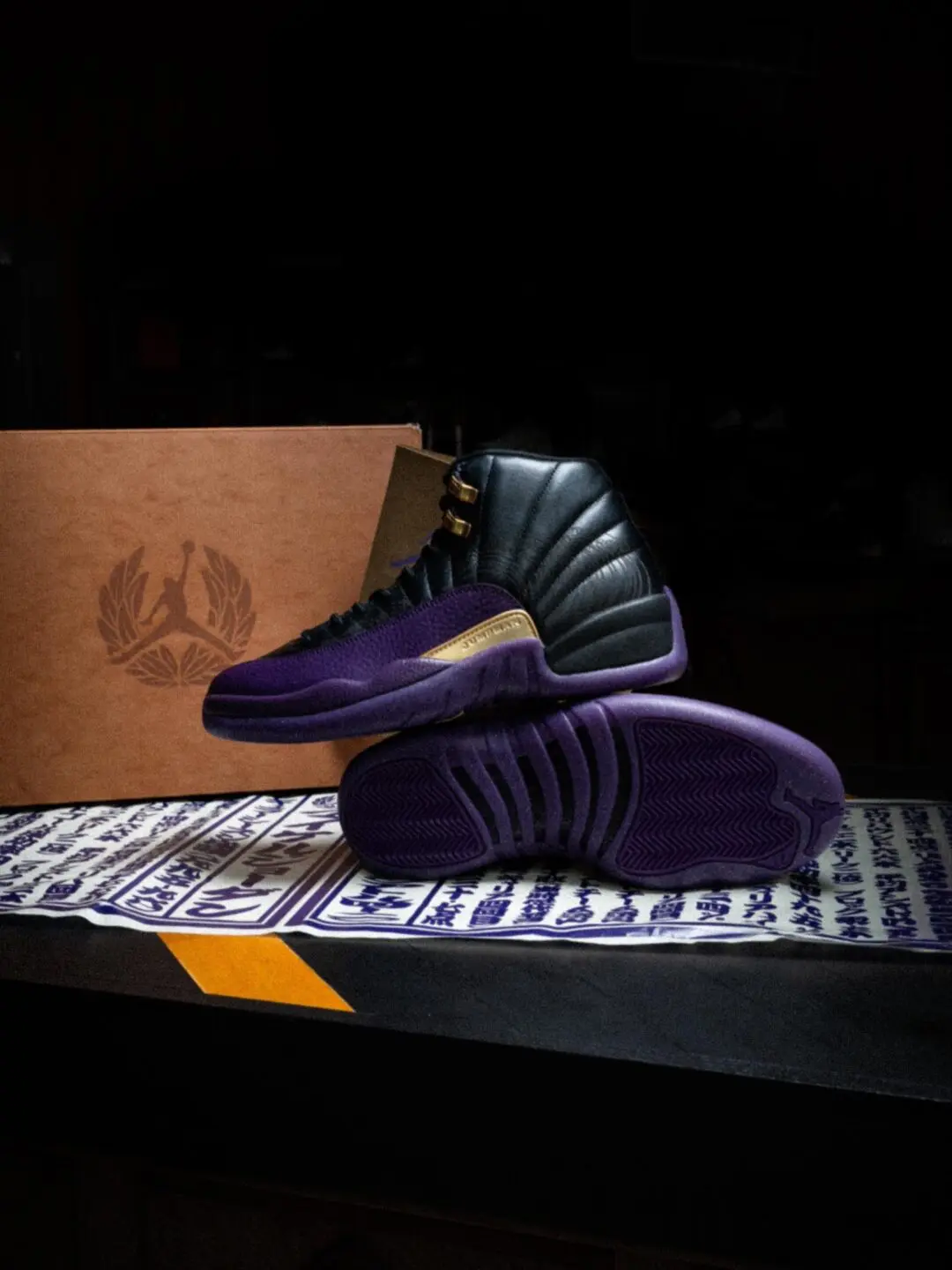 Pair Of Black And Deep Purple Air Jordan 12 Shoes Displayed With Gold Accents, Branded Brown Box, And Japanese-Inspired Graphic Paper.