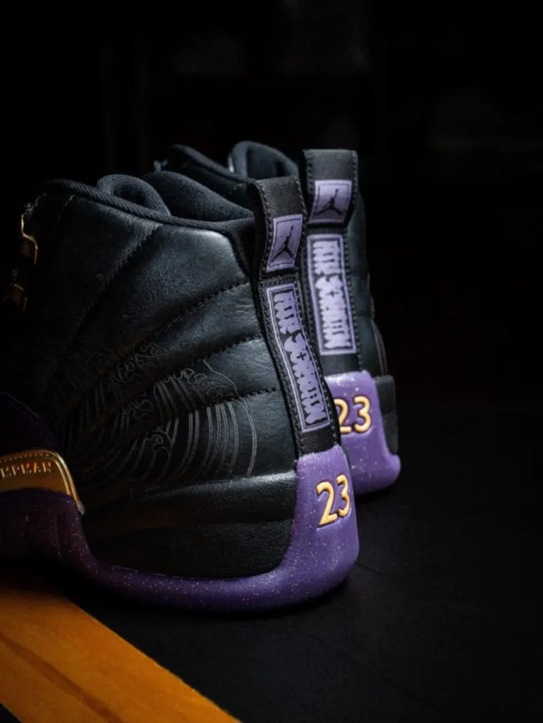 Rear Detail Of The Air Jordan 12 Show Black Leather Upper, Purple Heel Panel, Gold “23” Embroider, And Branded Pull Tab