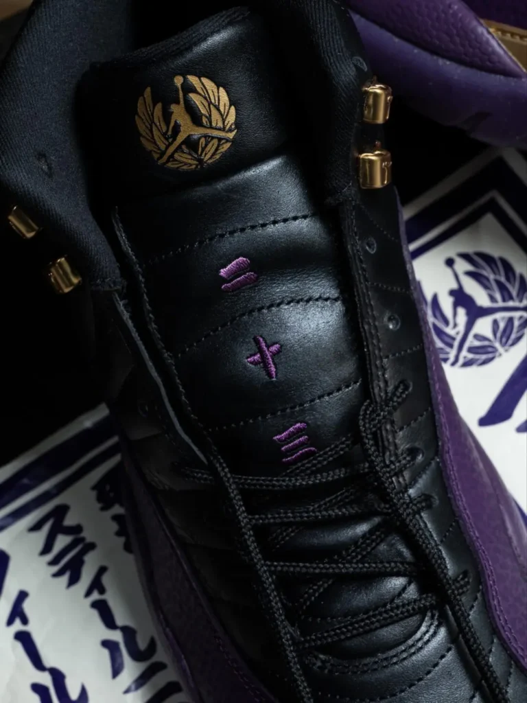 Close-Up Of The Air Jordan 12 Tongue Show A Gold Laurel Jumpman Emblem, Purple Embroider, Black Lace, And Gold Lace Hardware