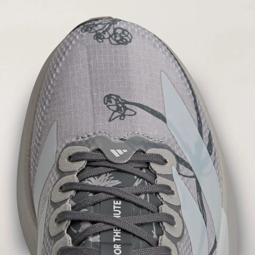 Close-up of the gray adidas Adizero shoe showing dark gray laces, pale three-stripe details, and black illustrated graphics across the toe.