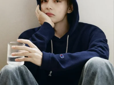 Person in an oversize navy Champion hoodie and faded wide-leg jean sits against a beige wall, rest their face on one hand while holding a glass of water