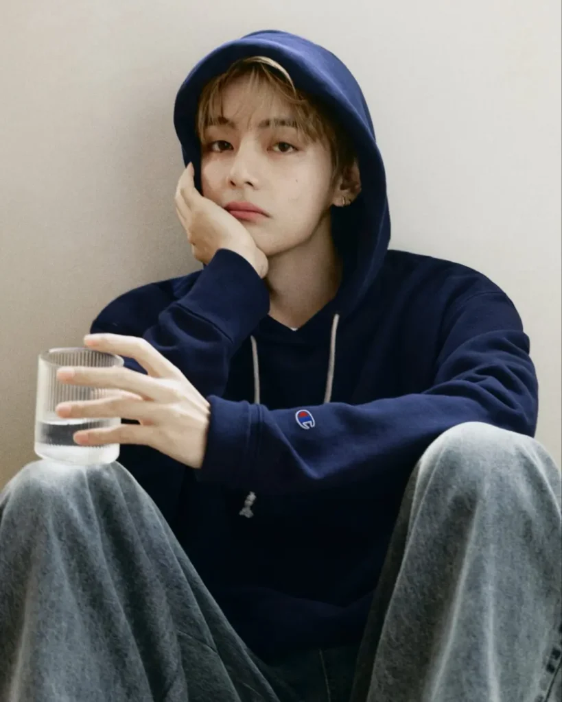Person in an oversize navy Champion hoodie and faded wide-leg jean sits against a beige wall, rest their face on one hand while holding a glass of water
