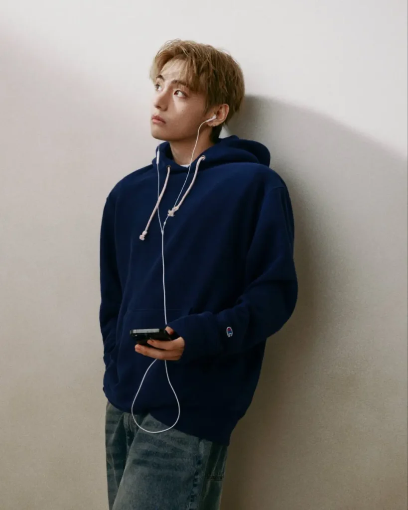 Person wearing a navy Champion hoodie and loose faded jeans leans against a plain wall, listening through white wired earphones while hold a smartphone