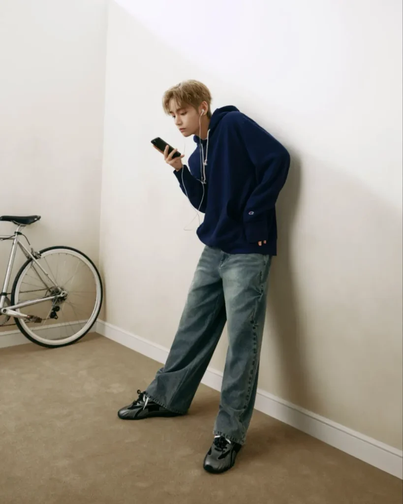 Person in an oversize navy Champion hoodie, faded wide-leg jeans and black shoes leans against a white wall while looking at a smartphone, with a silver bicycle position nearby