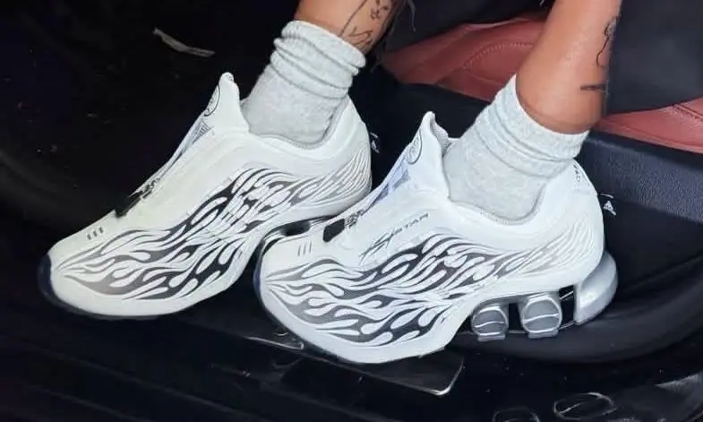 Close-up of white Hellstar x adidas shoes worn with gray socks, featuring black flame graphics and metallic silver heel cushioning.