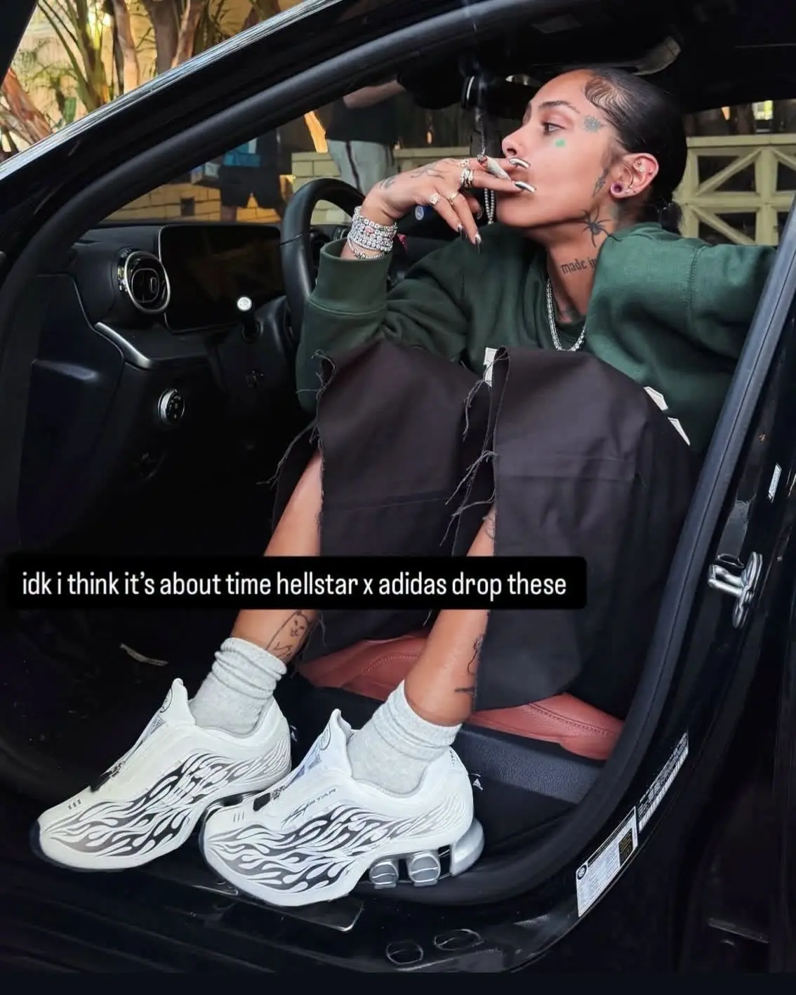 Person seated in a car wearing white Hellstar x adidas shoes with black flame graphics, zippered uppers, and metallic silver heel cushioning.