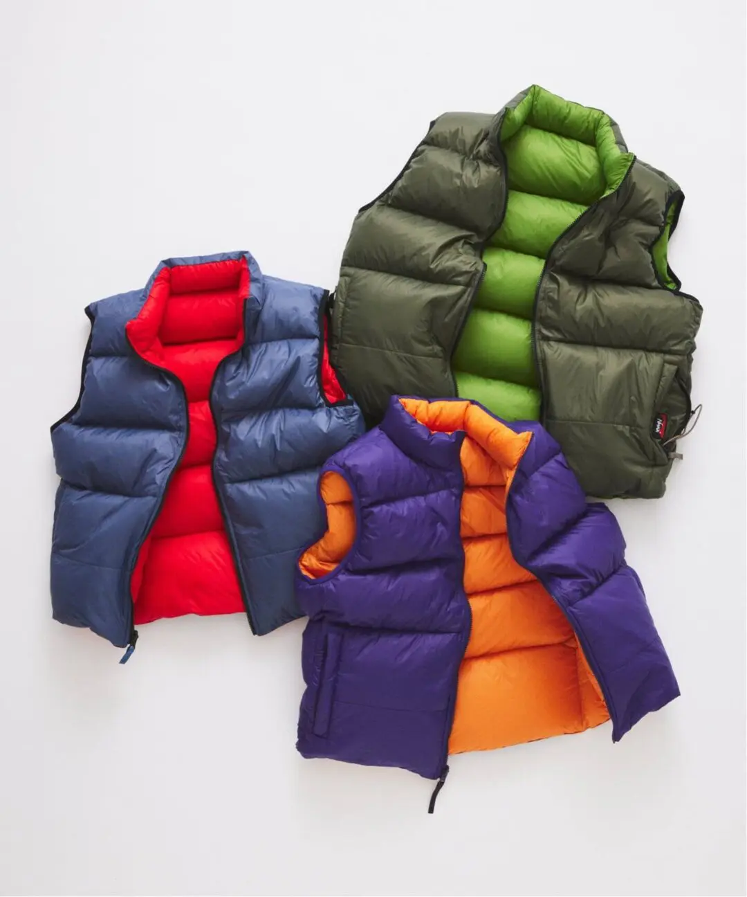 Three NANGA down vests on white: navy with red lining, olive with lime lining, purple with orange lining