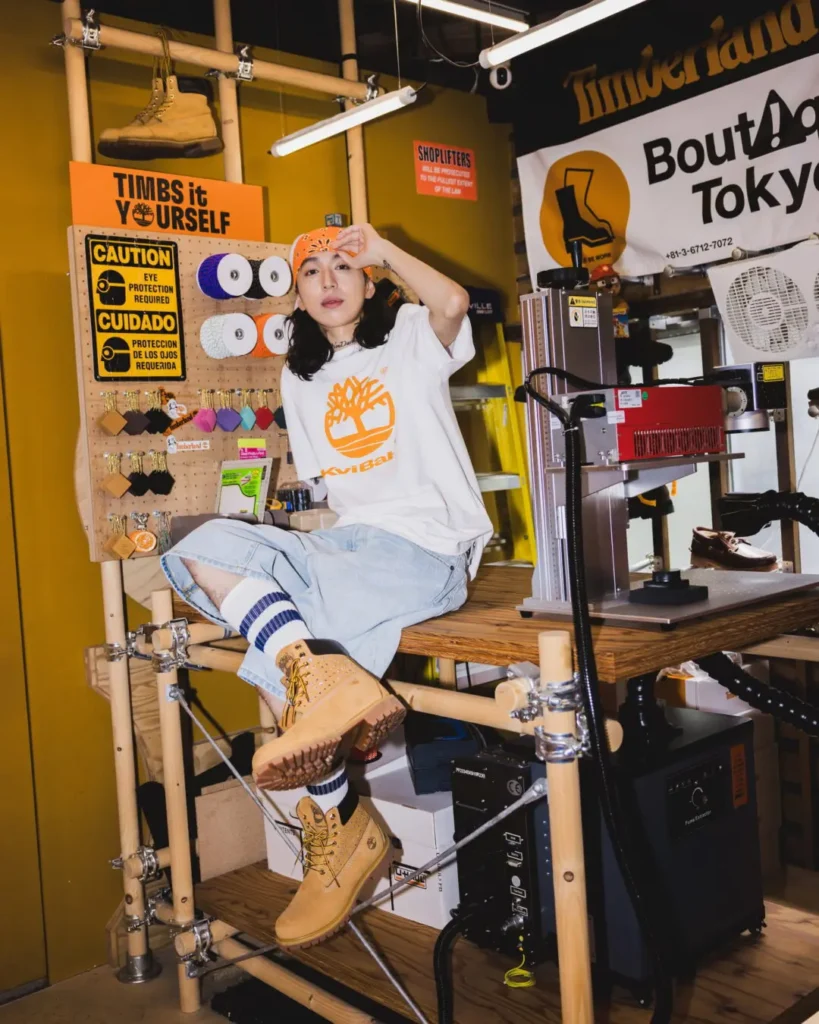 Model wear a white Timberland x Kvi Baba Tee, denim shorts, striped socks and wheat boot inside an industrial workshop