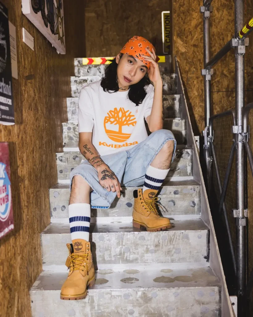 Model sit on metal stair wear an orange bandana, white graphic Tee, denim shorts and wheat Timberland boot