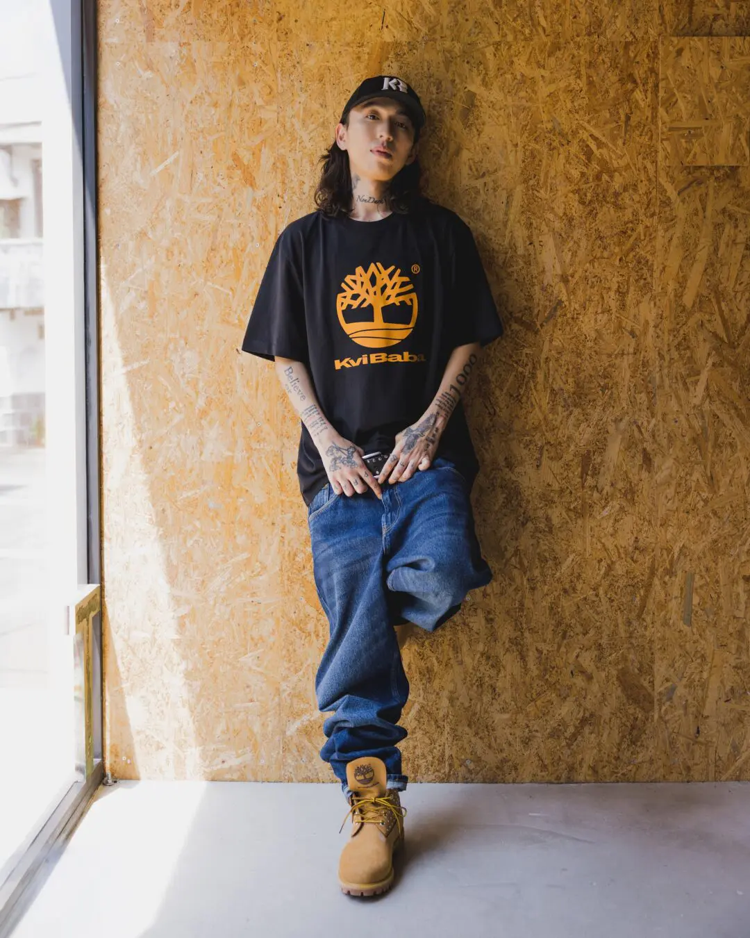 Kvi Baba Wearing A Black Timberland Collaboration T-Shirt, Blue Jeans And Wheat Timberland Boots While Posing Against A Plywood Wall.