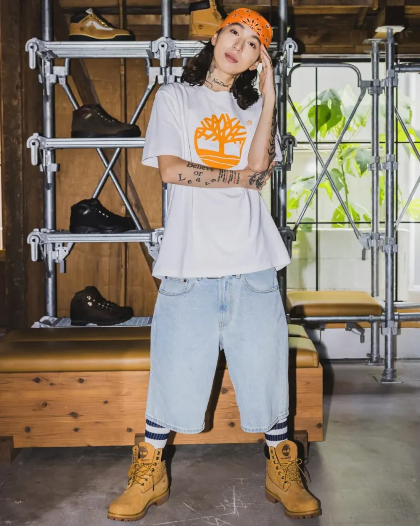 Model wear a white Timberland x Kvi Baba graphic Tee, denim short and wheat boot in front of industrial shelve