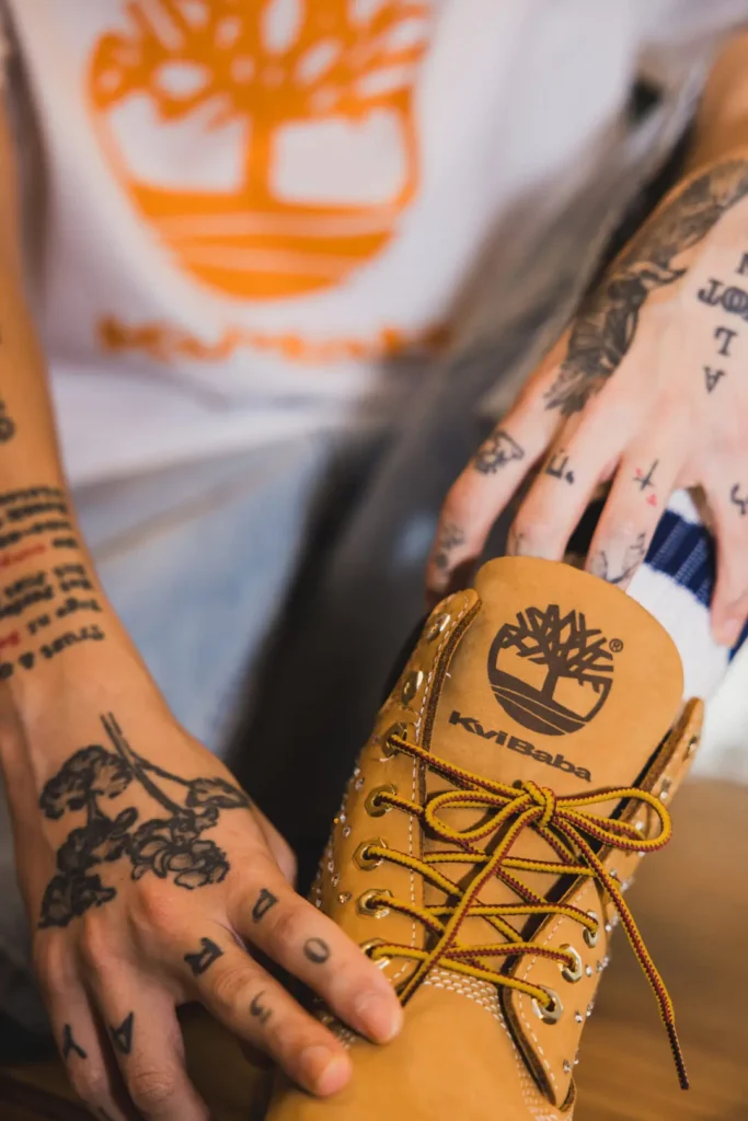 Close-up of tattooed hands adjust a wheat Timberland x Kvi Baba boot with brand tongue and two-tone lace