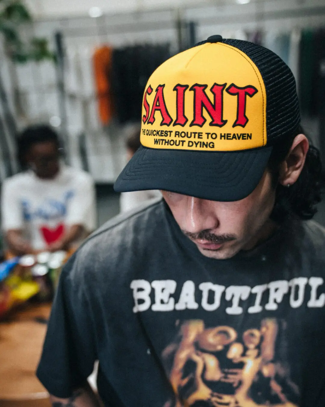 Person Wearing A Yellow And Black SAINT Trucker Cap With Red Gothic Branding And A Washed Dark Graphic T-Shirt.