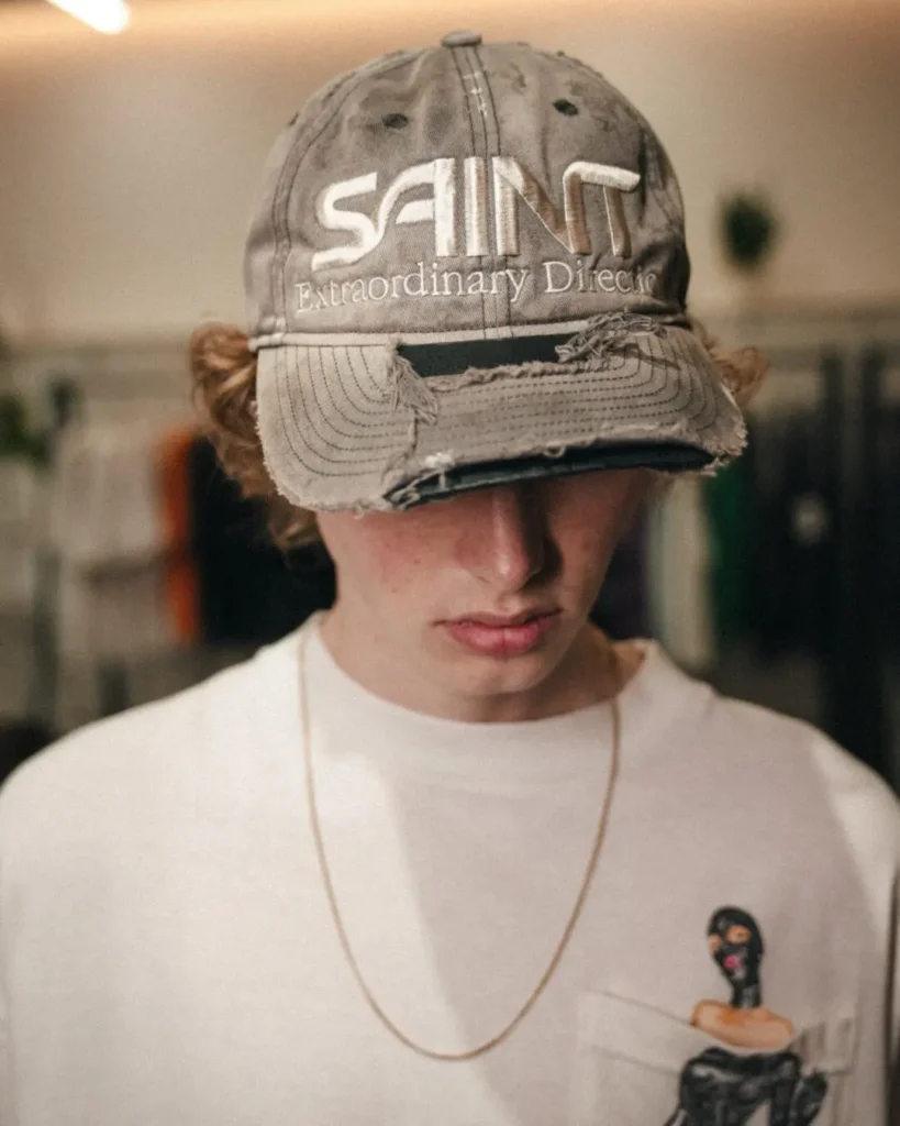 Person Wear A Distressed Gray SAINT Baseball Cap With White Embroider, Fray Brim, White Graphic Tee, And Gold-Tone Chain