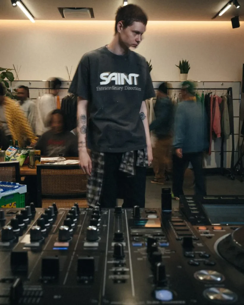 Person Wear A Washed Charcoal SAINT “Extraordinary Direction” Tee While Stand Behind DJ Equipment In A Clothing Store