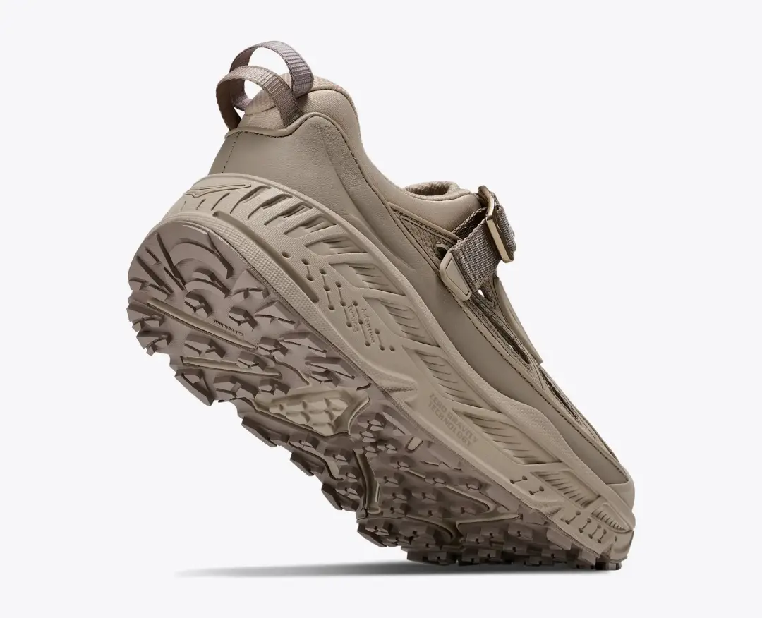 Rear three-quarter view of a beige HOKA trail shoe with an oversized cushioned midsole, rugged lug outsole, heel pull tab, and adjustable strap closure.