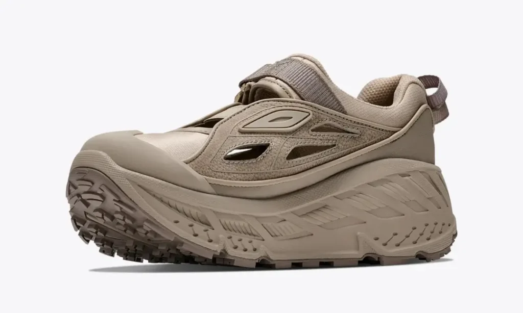 Front three-quarter view of a beige HOKA trail shoe highlighting the protective toe cap, open side ventilation, adjustable strap, and chunky trail sole