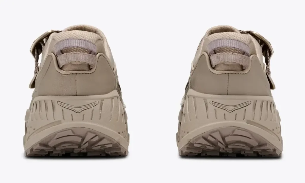 Rear view of a pair of beige HOKA trail shoes showing padded collars, heel pull tabs, sculpted midsoles, and aggressive lugged outsole